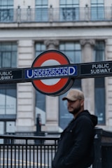 A man wearing a cap and dark jacket stands near a London Underground sign, with a blurred backdrop of a grand building featuring large windows and classical architecture.