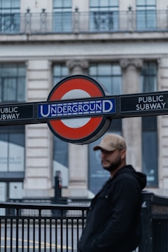 A man wearing a cap and dark jacket stands near a London Underground sign, with a blurred backdrop of a grand building featuring large windows and classical architecture.