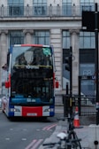 A double-decker bus with a Union Jack design on the front is on a city street, surrounded by urban architecture. Several passengers are visible on the top deck. A green sticker indicating 'Planet Friendly' is displayed on the windshield. The background includes a large building with columns and glass windows. There is a traffic light, street sign, and a bicycle on the side of the road.