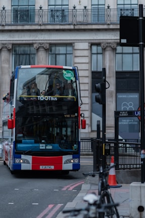 A double-decker bus with a Union Jack design on the front is on a city street, surrounded by urban architecture. Several passengers are visible on the top deck. A green sticker indicating 'Planet Friendly' is displayed on the windshield. The background includes a large building with columns and glass windows. There is a traffic light, street sign, and a bicycle on the side of the road.