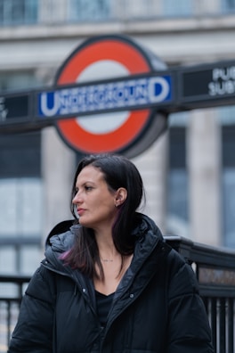 A woman in a black coat stands near an entrance to the London Underground. The iconic Underground sign with its red circle and blue background is prominently displayed in the background.