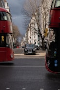 A black taxi drives down a city street lined with tall buildings. Red double-decker buses are visible on either side, framing the central taxi. The street is busy with traffic and pedestrians. The sky is overcast, adding a grey tone to the atmosphere. Leafless trees line the sidewalks.