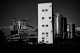 A black and white photograph of an industrial facility with large metal silos and a tall, rectangular corrugated metal building. Scaffolding and piping run across the scene, and there are clouds in the sky.