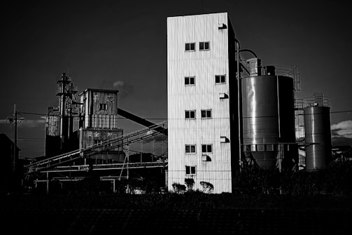 A black and white photograph of an industrial facility with large metal silos and a tall, rectangular corrugated metal building. Scaffolding and piping run across the scene, and there are clouds in the sky.