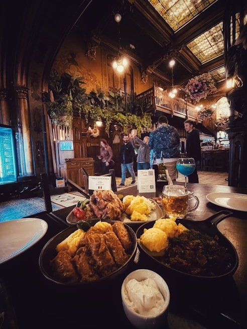 A richly decorated interior with ornate wooden architecture and lush greenery hanging from the ceiling. The room is dimly lit with warm lighting, creating a cozy atmosphere. In the foreground, a table is set with a variety of hearty dishes including meat, vegetables, and bread, accompanied by a mug of beer and a blue cocktail. People are gathered in the background, engaged in conversation near a staircase leading to a loft area.