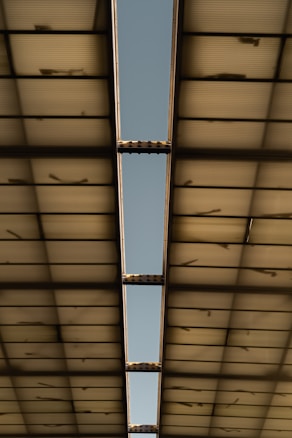A view of a metal structure with a long, narrow opening revealing a clear blue sky. The structure appears to be a roof or ceiling made of translucent panels supported by metallic beams.