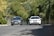 Two cars are driving side by side on a road bordered by lush green foliage. The road curves slightly, with a combination of sunlight and shadows creating a dynamic contrast on the asphalt. The surrounding environment is rich with greenery, providing a serene backdrop.