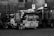 A black and white image of a row of graffiti-covered sanitation trucks parked closely together. The vehicles have various graffiti tags and designs on their sides. There is a pile of bags or refuse on top of one of the trucks. The trucks are situated in an urban area with a tall fence and a few visible signs.