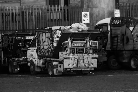 A black and white image of a row of graffiti-covered sanitation trucks parked closely together. The vehicles have various graffiti tags and designs on their sides. There is a pile of bags or refuse on top of one of the trucks. The trucks are situated in an urban area with a tall fence and a few visible signs.