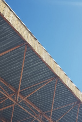 A view from below of a large industrial roof structure with exposed metal beams forming a grid pattern. The sky is clear and blue, creating a strong contrast with the earthy tones of the beams.