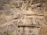 A close-up view of a rocky cliff face with layers of sedimentary rock. The surface appears weathered and eroded, showing various shades of brown and gray. Some areas have cracks and small protrusions, indicating natural aging and the effects of environmental factors.