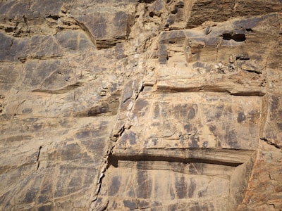A close-up view of a rocky cliff face with layers of sedimentary rock. The surface appears weathered and eroded, showing various shades of brown and gray. Some areas have cracks and small protrusions, indicating natural aging and the effects of environmental factors.