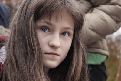 A young girl with long brown hair and blue eyes looks directly at the camera with a serious expression. She is wearing a light-colored coat. In the background, there is a person wearing a padded jacket.