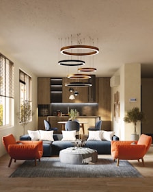 A modern living room with a stylish interior design, featuring a blue sectional sofa adorned with white cushions, an orange armchair, and a round gray pouf on a textured rug. Modern circular chandeliers hang from the ceiling above the seating area. The back of the room showcases a small dining area with chairs and a wooden kitchen setup. Large windows on the left side allow natural light to flood the space, highlighting the wooden flooring and potted trees placed at the corners.