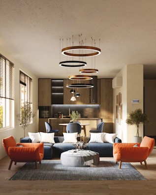 A modern living room with a stylish interior design, featuring a blue sectional sofa adorned with white cushions, an orange armchair, and a round gray pouf on a textured rug. Modern circular chandeliers hang from the ceiling above the seating area. The back of the room showcases a small dining area with chairs and a wooden kitchen setup. Large windows on the left side allow natural light to flood the space, highlighting the wooden flooring and potted trees placed at the corners.