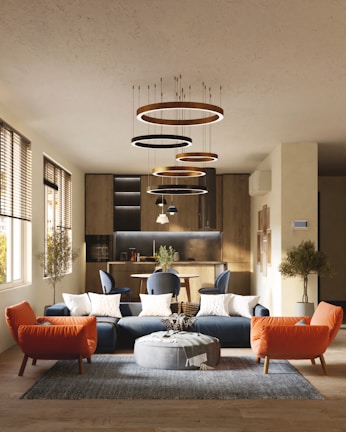 A modern living room with a stylish interior design, featuring a blue sectional sofa adorned with white cushions, an orange armchair, and a round gray pouf on a textured rug. Modern circular chandeliers hang from the ceiling above the seating area. The back of the room showcases a small dining area with chairs and a wooden kitchen setup. Large windows on the left side allow natural light to flood the space, highlighting the wooden flooring and potted trees placed at the corners.