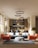 A modern living room with a stylish interior design, featuring a blue sectional sofa adorned with white cushions, an orange armchair, and a round gray pouf on a textured rug. Modern circular chandeliers hang from the ceiling above the seating area. The back of the room showcases a small dining area with chairs and a wooden kitchen setup. Large windows on the left side allow natural light to flood the space, highlighting the wooden flooring and potted trees placed at the corners.