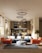 A modern living room with a stylish interior design, featuring a blue sectional sofa adorned with white cushions, an orange armchair, and a round gray pouf on a textured rug. Modern circular chandeliers hang from the ceiling above the seating area. The back of the room showcases a small dining area with chairs and a wooden kitchen setup. Large windows on the left side allow natural light to flood the space, highlighting the wooden flooring and potted trees placed at the corners.