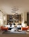 A modern living room with a stylish interior design, featuring a blue sectional sofa adorned with white cushions, an orange armchair, and a round gray pouf on a textured rug. Modern circular chandeliers hang from the ceiling above the seating area. The back of the room showcases a small dining area with chairs and a wooden kitchen setup. Large windows on the left side allow natural light to flood the space, highlighting the wooden flooring and potted trees placed at the corners.