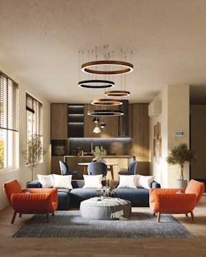 A modern living room with a stylish interior design, featuring a blue sectional sofa adorned with white cushions, an orange armchair, and a round gray pouf on a textured rug. Modern circular chandeliers hang from the ceiling above the seating area. The back of the room showcases a small dining area with chairs and a wooden kitchen setup. Large windows on the left side allow natural light to flood the space, highlighting the wooden flooring and potted trees placed at the corners.