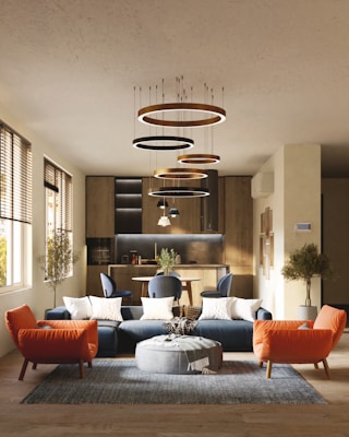 A modern living room with a stylish interior design, featuring a blue sectional sofa adorned with white cushions, an orange armchair, and a round gray pouf on a textured rug. Modern circular chandeliers hang from the ceiling above the seating area. The back of the room showcases a small dining area with chairs and a wooden kitchen setup. Large windows on the left side allow natural light to flood the space, highlighting the wooden flooring and potted trees placed at the corners.