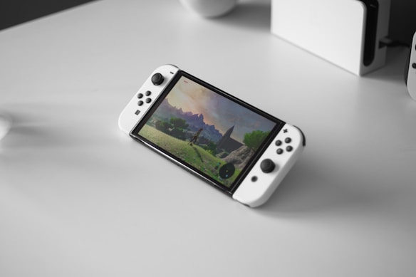 A portable gaming console with a screen showing a video game landscape, resting on a light-colored surface. The console has white controllers attached to either side of the screen. Nearby, there seems to be a white docking station or accessory.