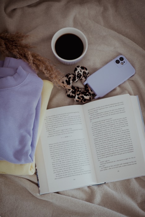 An open book rests on a soft, beige fabric surface surrounded by items such as a lavender sweater, a yellow garment, a cup of black coffee, a purple phone case, and a leopard print scrunchie. Dried flowers add a delicate, natural touch.