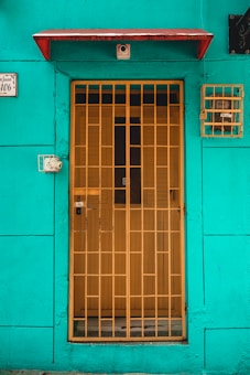 A vibrant turquoise wall with a yellow metal gate covering a wooden door. Above the door is a small red awning, and there is a security camera installed just below it. On the left side, a small black and white sign displays the number 106. To the right of the door, a small yellow metal cage encloses a utility meter.