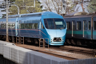 A sleek blue high-speed passenger train travels on a railway track amidst a backdrop of trees and overhead electrical lines. The train is prominent and appears to be moving, with its modern aerodynamic design and smooth finish. Another train with a similar color scheme is seen alongside it, emphasizing a busy rail system.
