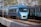 A sleek blue high-speed passenger train travels on a railway track amidst a backdrop of trees and overhead electrical lines. The train is prominent and appears to be moving, with its modern aerodynamic design and smooth finish. Another train with a similar color scheme is seen alongside it, emphasizing a busy rail system.
