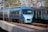 A sleek blue high-speed passenger train travels on a railway track amidst a backdrop of trees and overhead electrical lines. The train is prominent and appears to be moving, with its modern aerodynamic design and smooth finish. Another train with a similar color scheme is seen alongside it, emphasizing a busy rail system.