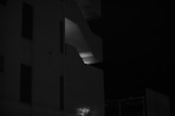 A dark and moody black and white photograph focusing on a modern building's architectural elements. The structure features prominent lines and geometric shapes, with a single illuminated section around a stairwell, creating a strong contrast. The darkness of the scene enhances the overall mysterious ambiance.