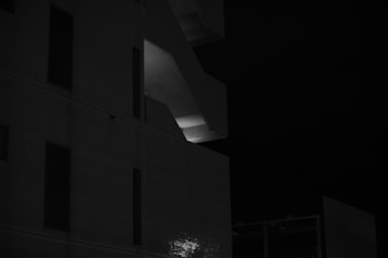 A dark and moody black and white photograph focusing on a modern building's architectural elements. The structure features prominent lines and geometric shapes, with a single illuminated section around a stairwell, creating a strong contrast. The darkness of the scene enhances the overall mysterious ambiance.