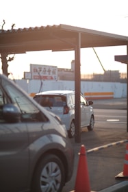 Two cars are parked under a metal carport, with one vehicle predominantly silver and another white. An orange traffic cone is visible in the foreground. The background displays a billboard with Japanese text and a partially visible construction site, under warm sunlight.