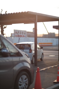 Two cars are parked under a metal carport, with one vehicle predominantly silver and another white. An orange traffic cone is visible in the foreground. The background displays a billboard with Japanese text and a partially visible construction site, under warm sunlight.