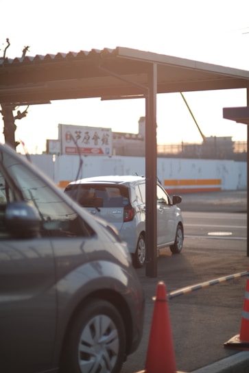 Two cars are parked under a metal carport, with one vehicle predominantly silver and another white. An orange traffic cone is visible in the foreground. The background displays a billboard with Japanese text and a partially visible construction site, under warm sunlight.