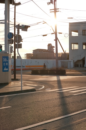 Sunlight shines through an urban street intersection with traffic lights and road signs. Power lines crisscross above, and there is a distant building with a clear view of the sky.