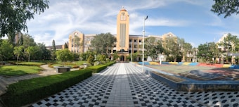 A grand, symmetrical building with a clock tower in the center, surrounded by a manicured garden with green trees and shrubs. The foreground features a black and white tiled pathway leading towards the building, flanked by well-maintained lawns. A colorful sports court is visible on the right side.