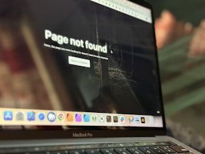 A laptop displays a 'Page not found' message on its screen, indicating a missing webpage. The screen shows a dimly lit background, possibly resembling an indoor setting. The laptop sits at an angle, with the keyboard and trackpad partially visible. A row of application icons is present at the bottom of the screen.