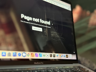 A laptop displays a 'Page not found' message on its screen, indicating a missing webpage. The screen shows a dimly lit background, possibly resembling an indoor setting. The laptop sits at an angle, with the keyboard and trackpad partially visible. A row of application icons is present at the bottom of the screen.