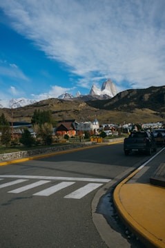 A scenic view of a quiet town with a road leading towards distant mountains. The sky is partly cloudy with a vivid blue hue, and the mountains in the background are capped with snow. Houses with colorful roofs are nestled between greenery, and vehicles are parked along the road.