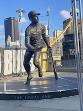 A bronze statue of a baseball player is captured in mid-stride with a bat in hand, set against a backdrop of city skyscrapers and a bridge. The statue stands on a pedestal inscribed with 'The Great One.' Surrounding the statue are lamp posts and a partially visible banner.