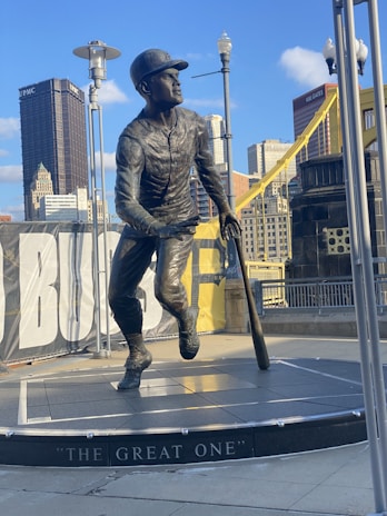 A bronze statue of a baseball player is captured in mid-stride with a bat in hand, set against a backdrop of city skyscrapers and a bridge. The statue stands on a pedestal inscribed with 'The Great One.' Surrounding the statue are lamp posts and a partially visible banner.