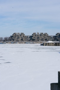 A snow-covered landscape with a unique cluster of modular concrete buildings in the background. The buildings have a distinctive, repetitive design. In the foreground, there is a large ice-covered body of water, possibly a lake or river. The sky is mostly clear with some light clouds.