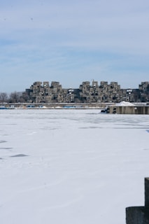 A snow-covered landscape with a unique cluster of modular concrete buildings in the background. The buildings have a distinctive, repetitive design. In the foreground, there is a large ice-covered body of water, possibly a lake or river. The sky is mostly clear with some light clouds.