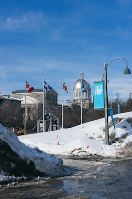 Flags from Canada and Quebec flutter next to a historic building with a prominent dome. Snow-covered ground and piles of snow are visible, indicating a winter setting. A sign reads 'Old Port,' suggesting the location.