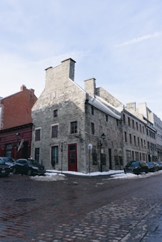 A historic stone building with red-framed windows and a red door stands at a street corner. The structure is surrounded by cobblestone streets and patches of snow, with several parked cars nearby. The sky is clear with a few clouds, and the area has street lamps and other architectural features suggesting an old town or heritage district.