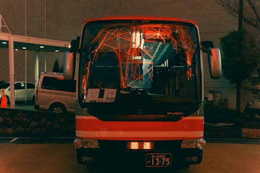A bus is parked in an urban setting, illuminated by various lights that cast an array of orange and red reflections on its windshield. The bus has a black and red color scheme and displays a license plate. There are other vehicles parked nearby and a dimly lit building in the background.