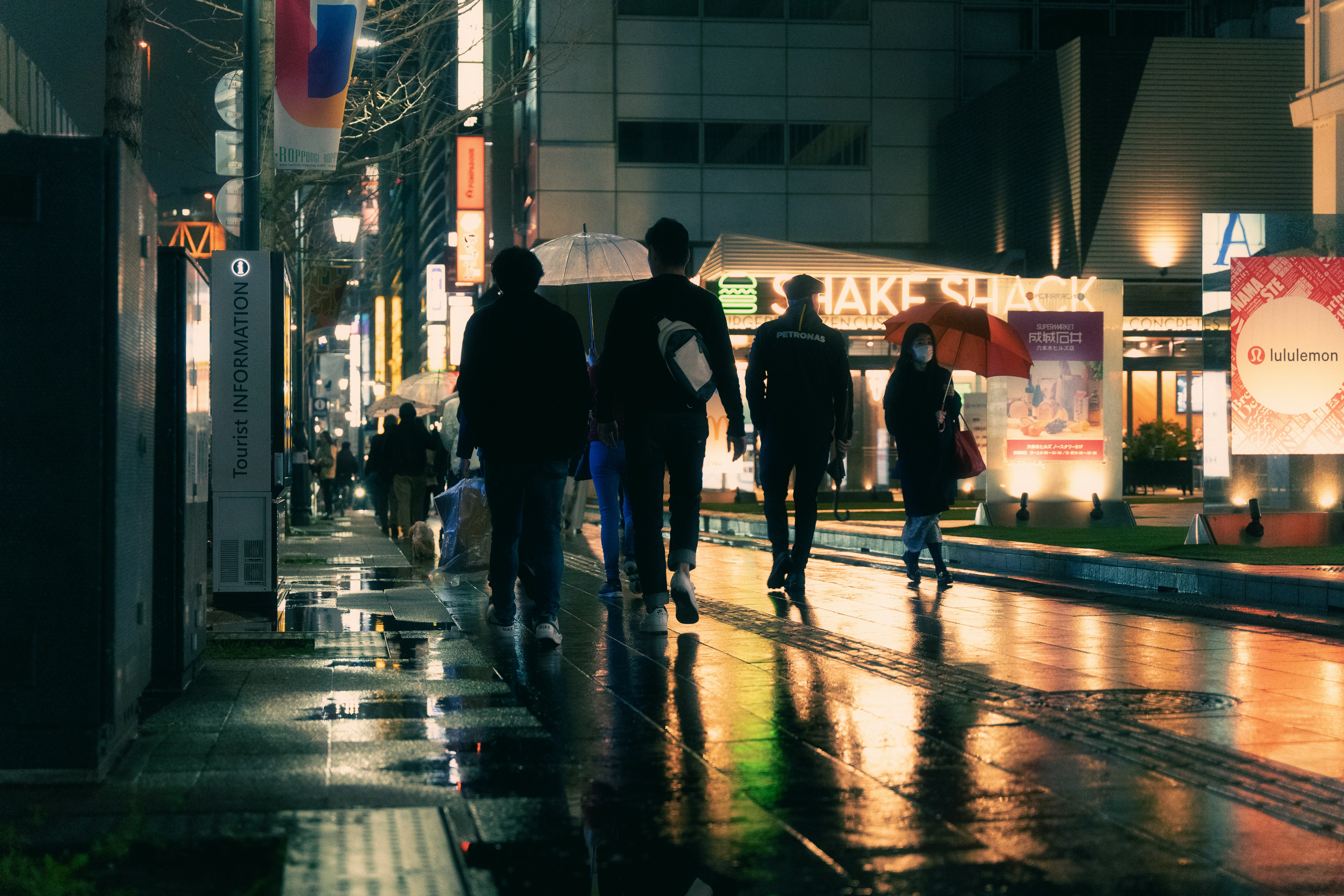 Night time, city scene. People in silhouette walk down a city street, lights from shops are reflected in the wet road.