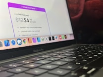 A close-up view of a MacBook Pro showing a section of the screen and keyboard. The screen displays a marketing offer for a subscription service with a limited launch special price. Various app icons are visible on the dock at the bottom of the screen.
